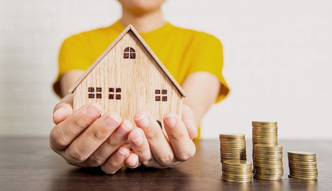 Frau hält ein kleines Haus aus Holz, daneben stehen 3 Stapel Münzen auf dem Tisch